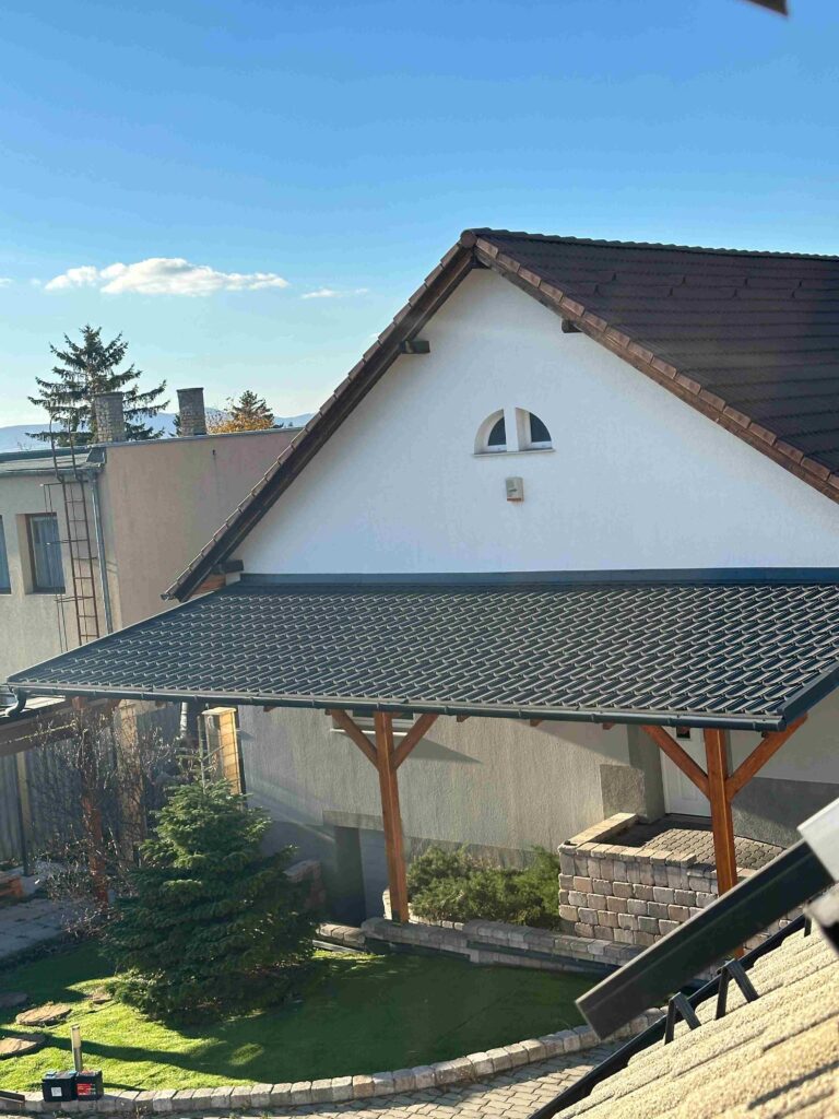 A black, tile-patterned roof covers a timber-supported carport in the foreground. Behind it, a white gabled building has a dark reddish-brown roof and a small arched upper window. Blue sky, trees, paved ground, and stacks of masonry blocks surround the structures.