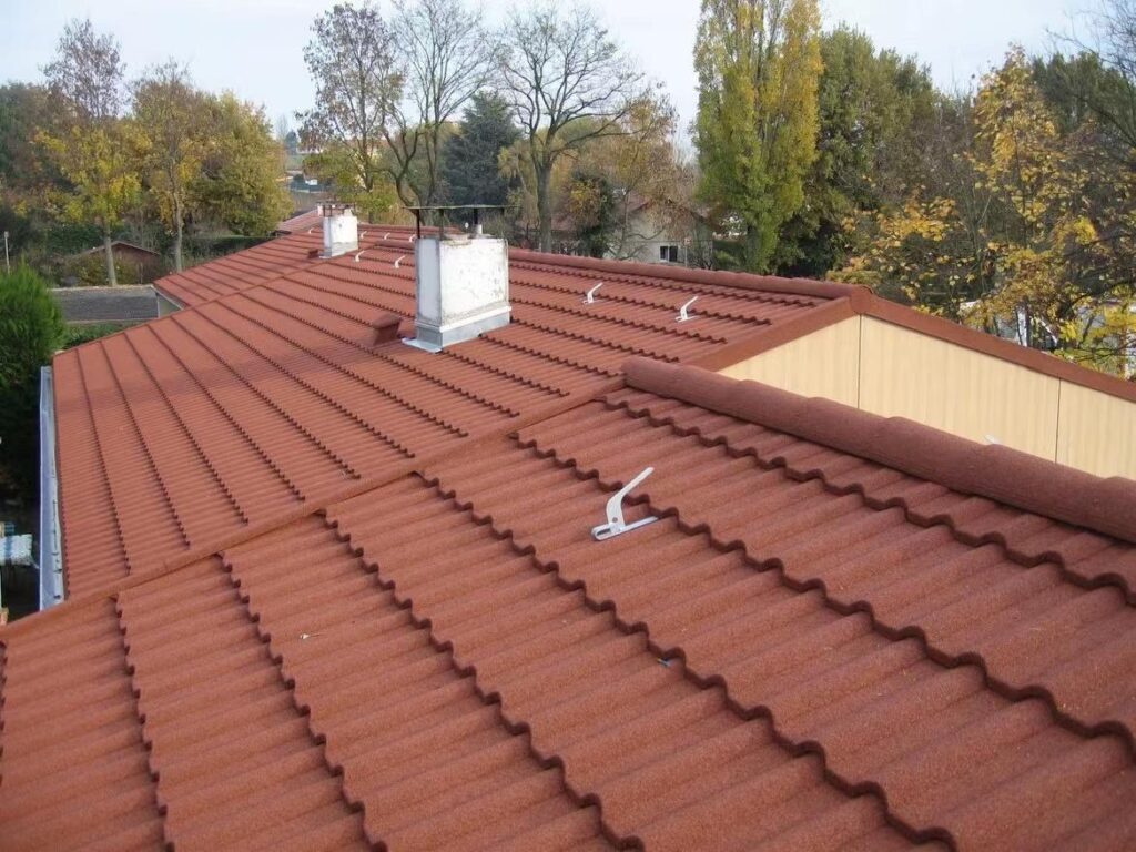 A broad finished roof fills most of the image, with orderly rows of reddish-brown profiled tiles across multiple roof planes. Several white chimneys and small white roof fixtures project above the surface. Trees with autumn-colored foliage and neighboring rooflines appear in the distance.
