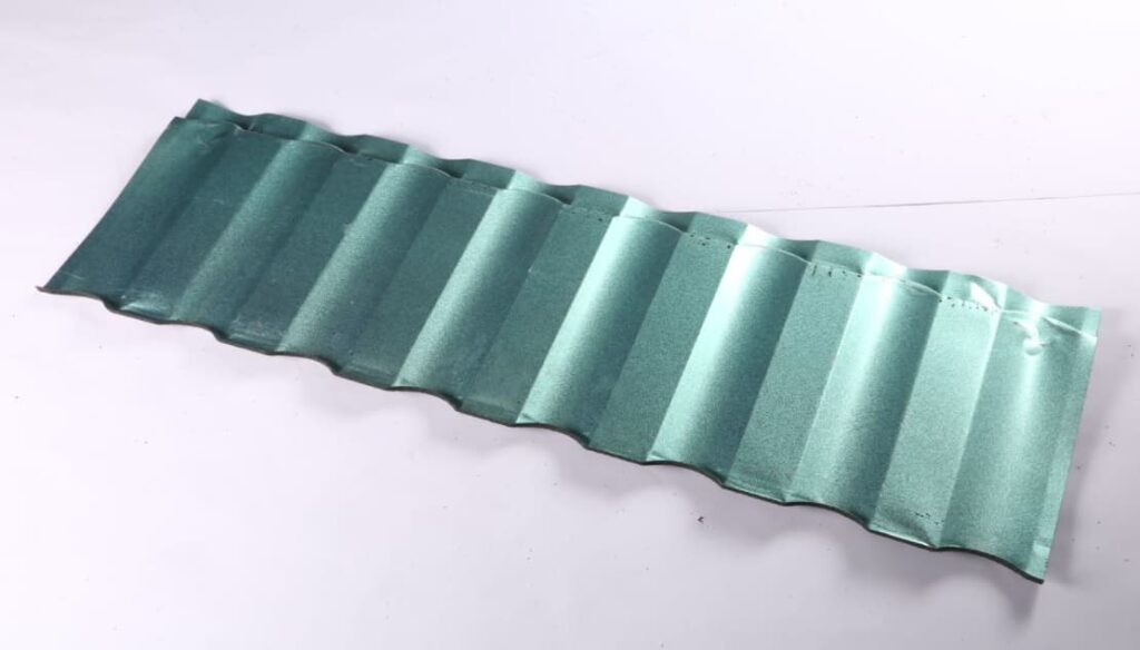 A single long green-gray roofing panel lies diagonally on a plain light background. Its surface has repeated raised ribs and shallow troughs running across the width. Small dark marks and slight bends are visible near parts of its edges.
