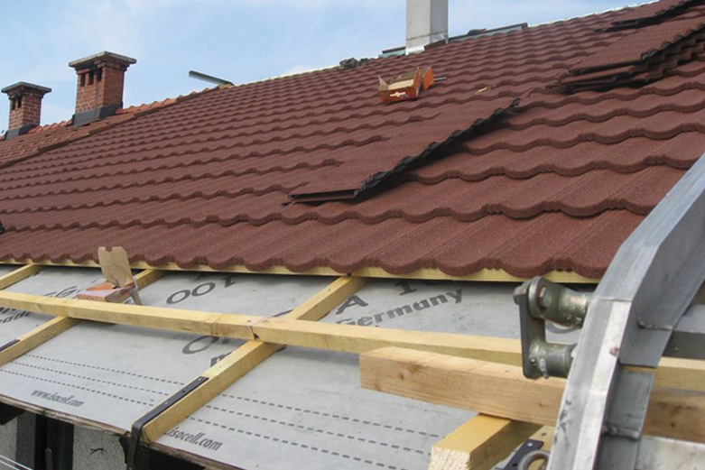The image shows a sloping roof partly covered with reddish-brown wavy panels. The lower portion remains open, exposing pale membrane and wooden battens, with partial printed markings on the membrane and a metal lifting fixture at the right edge. Chimneys and a strip of pale sky are visible above the roof.