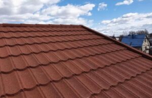 An oblique view shows a broad red-brown pitched roof made of repeating curved tile-like courses. A roof ridge runs horizontally near the upper part of the frame, with pale blue clouds behind it. Part of a neighboring blue roof and roofline is visible at the upper right.