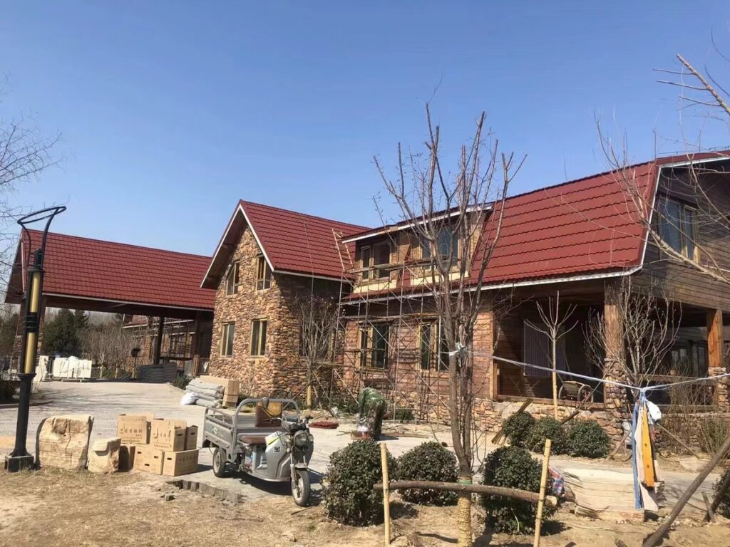 A large stone-faced building with multiple steep red-brown tiled roof sections fills the scene. Scaffolding is set along parts of the facade, and an open porch-like area appears at the right. Boxes, masonry pieces, equipment, and other materials occupy the paved foreground beneath a clear blue sky.