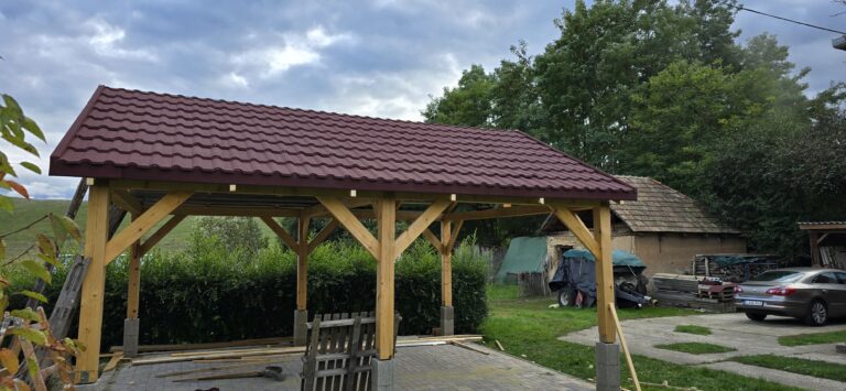 An open-sided timber shelter fills the foreground, with wooden posts and braces supporting a broad red-brown roof of repeating tile-like courses. The shelter stands on a paved area edged by low walls and dense shrubs. Trees, a cloudy sky, a parked car, and other structures appear behind it.