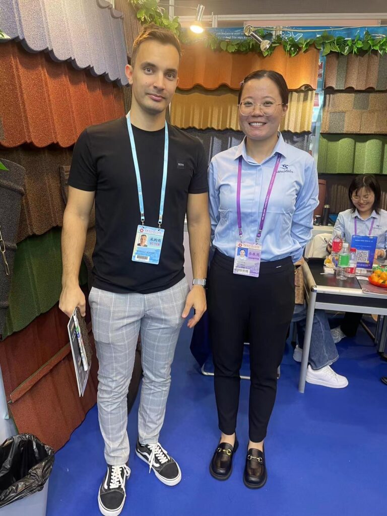 A vertical indoor photo shows two adults standing in front of wall-mounted roofing samples in several colors and profiles. Both wear lanyards with ID badges; the person on the left wears a black shirt and light gray plaid trousers, while the person on the right wears glasses and a light shirt. A seated person, leafy decorations, and a table with small items are partially visible on the right.