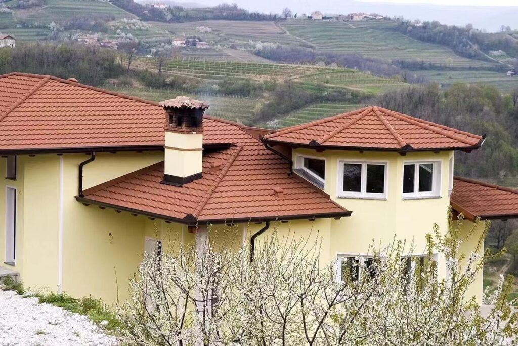 A pale yellow multi-section house fills the foreground, with broad red-orange tiled roofs and a tall chimney. Dark gutters trace the eaves, and white-framed windows sit below a projecting roof section. Terraced rows and scattered buildings cover the distant hillside behind the house.