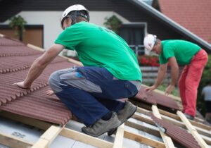 A helmeted person in a green shirt and dark blue trousers crouches on a sloped roof covered with reddish-brown panels. Another helmeted person bends over farther along the roof, and pale membrane with timber supports is visible at the bottom edge. A dark house facade and nearby roof surfaces appear in the background.