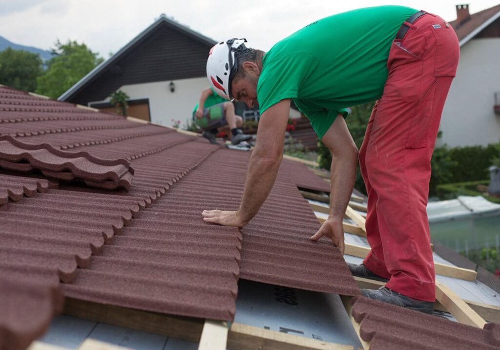A person wearing a white helmet, green shirt, and red trousers is bent over a sloped roof. Both hands rest on a reddish-brown profiled panel positioned among similar panels. Pale membrane and timber supports are exposed at the lower edge, with buildings visible behind.