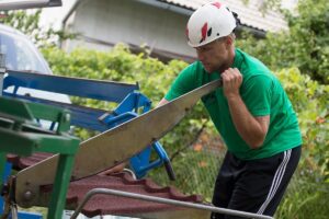 A helmeted person in a green shirt and dark trousers stands beside a blue-framed machine and handles a long flat metal piece held diagonally across the body. Reddish-brown profiled panels are visible in the foreground beneath the machine. Green foliage and a building exterior form the background.