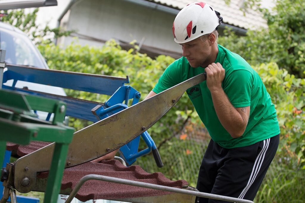 A helmeted person in a green shirt and dark trousers stands beside a blue-framed machine on a roof. They hold a long flat metal piece at the machine, with reddish-brown profiled panels visible below and behind it. Foliage and a pale roof surface fill the background.