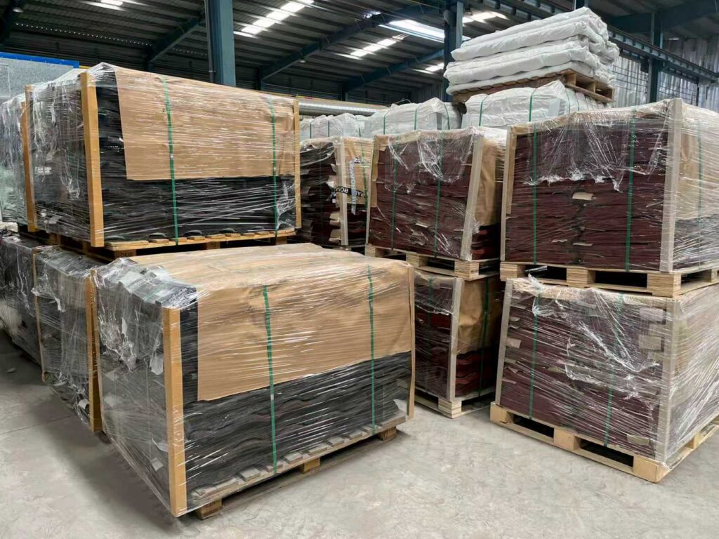 Several large rectangular loads rest on wooden pallets inside a roofed storage space. Clear plastic film and green bands secure the loads; some faces appear dark while others are partly covered with tan sheets. Stacks of white bagged material and more wrapped loads are visible behind them.
